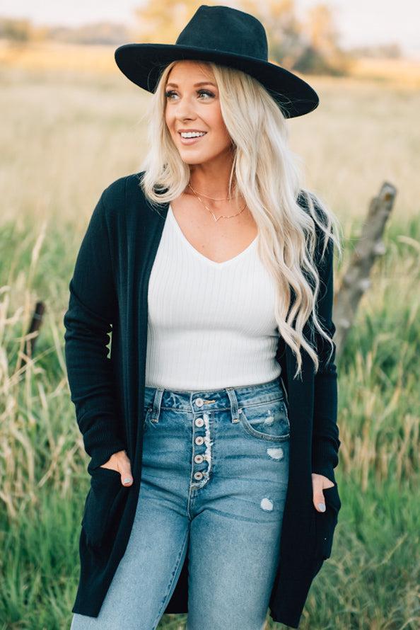 A woman wearing a long black cardigan with pockets, paired with a white top and blue jeans, standing in a field.