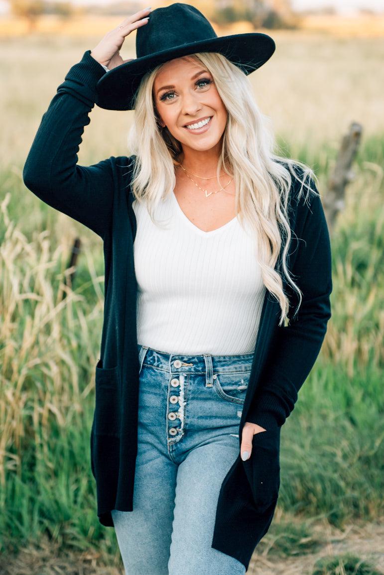 A woman wearing a long cardigan with pockets, paired with a white top and blue jeans, standing in a field.