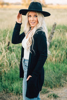 A woman wearing a long black cardigan with pockets, paired with a white top and blue jeans, standing in a field.