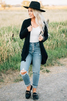 A woman wearing a long cardigan with pockets, paired with a white top and blue jeans, standing in a field.