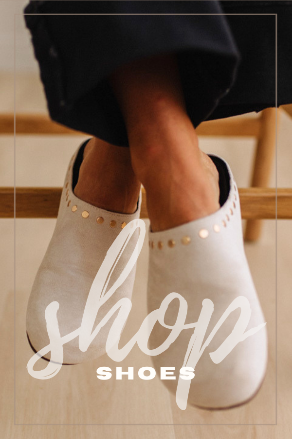 A close-up lifestyle shot of a person wearing light taupe studded mule shoes, with dark pants and a wooden chair visible in the background. A large white script overlay reads 'shop' and 'SHOES' is printed below.