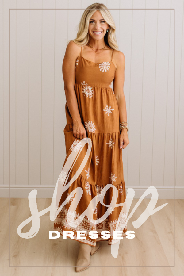 A woman modeling a camel colored tiered maxi dress with white floral embroidery, standing against a white paneled wall. A large white script overlay reading 'shop' and 'DRESSES' is across the lower portion of the image.