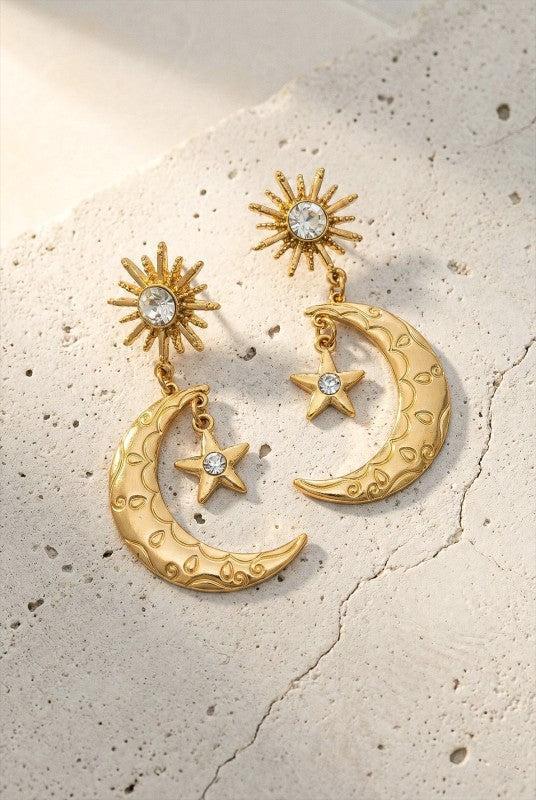 Gold earrings with moon, star, and sun designs on a textured beige surface