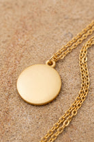 A gold disc pendant necklace with a matching chain, displayed on a textured beige sand-like surface.