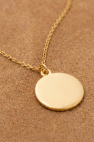 A gold disc pendant necklace on a matching gold chain, displayed on a textured brown background.