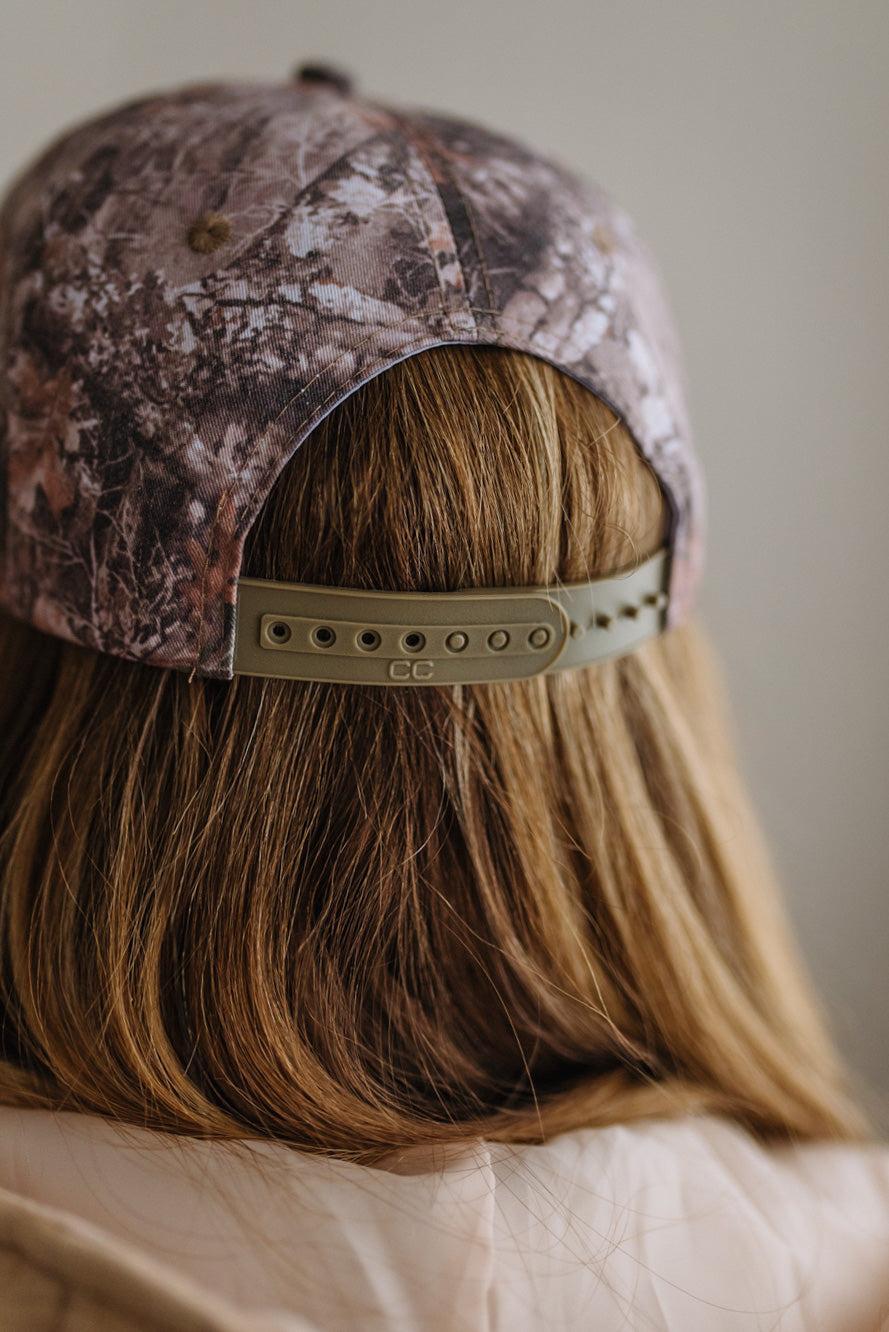 Close-up back view of a person wearing a camouflage baseball cap with a plastic adjustable strap and a light brown hair.