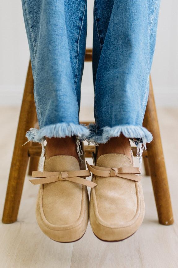 A person sitting on a wooden stool wearing beige suede slip-on loafers with bow details and frayed blue jeans.