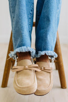 A person sitting on a wooden stool wearing beige suede slip-on loafers with bow details and frayed blue jeans.