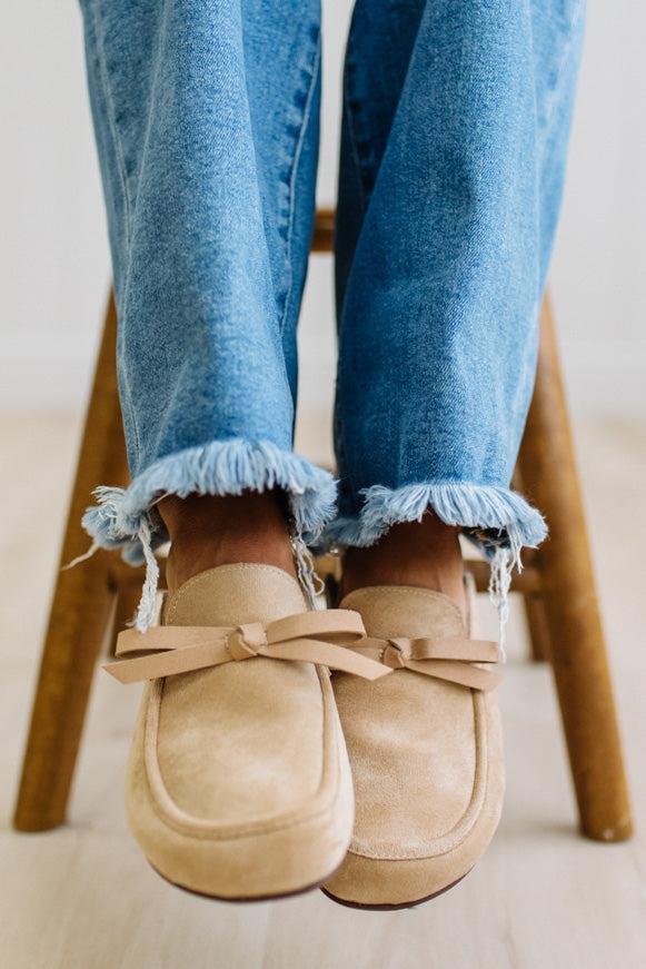 A person sitting on a wooden stool wearing beige suede slip-on loafers with bow details and frayed blue jeans.