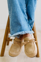 A person sitting on a wooden stool wearing beige suede slip-on loafers with bow details and frayed blue jeans.
