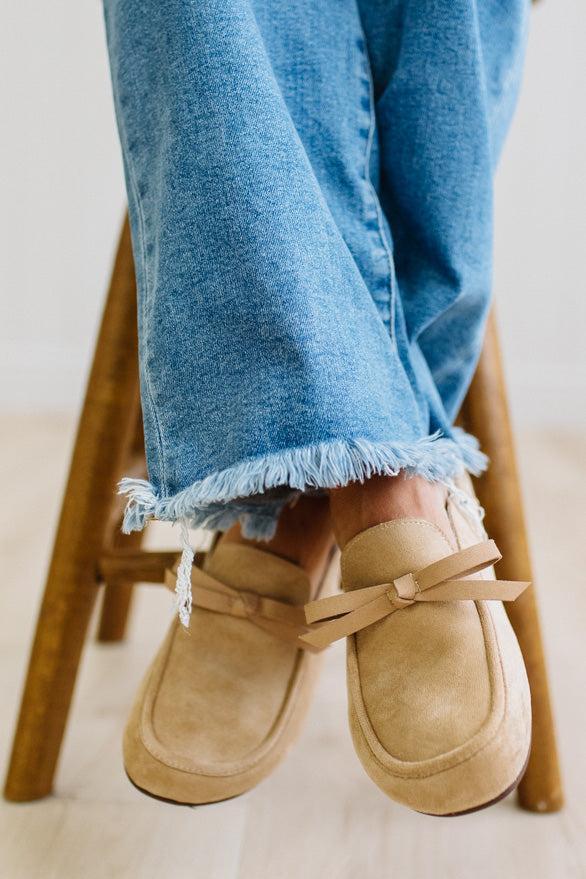A person sitting on a wooden stool wearing beige suede slip-on loafers with bow details and frayed blue jeans.