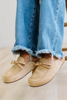 A person sitting on a wooden stool wearing beige suede slip-on loafers with bow details and frayed blue jeans.
