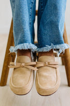 A close-up of a person's feet wearing beige suede loafers with matching ribbon bows, paired with frayed blue jeans, sitting on a wooden stool.