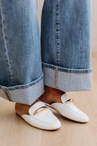 White loafers with gold accents worn with rolled-up blue jeans on a neutral background