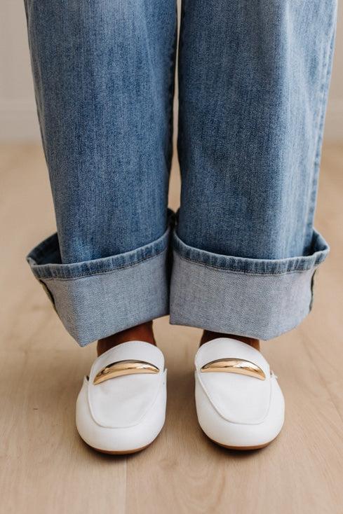 White loafers with gold accents worn with rolled-up blue jeans on a wooden floor.