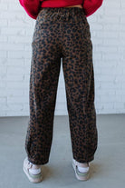 Person wearing leopard print pants with a red sweater and white sneakers against a white brick wall.