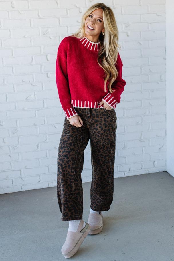 Woman wearing a red sweater and leopard print pants against a white brick wall.