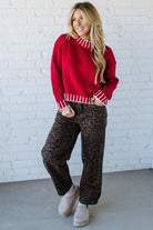 Woman wearing a red sweater and leopard print pants against a white brick wall.