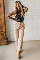 Woman wearing a floral top and beige pants against a white brick wall.