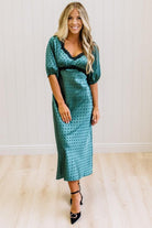 A woman modeling a teal green midi-length slip dress with a black polka dot pattern, black lace trim at the V-neck, and puff sleeves, paired with black heels, standing in a room with white paneled walls and light wood floors.