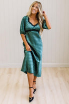 A woman modeling a teal green midi-length slip dress with a black polka dot pattern, black lace trim at the V-neck, and puff sleeves, paired with black heels, standing in a room with white paneled walls and light wood floors.