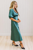 A woman modeling a teal green midi-length slip dress with a black polka dot pattern, black lace trim at the V-neck, and puff sleeves, paired with black heels, standing in a room with white paneled walls and light wood floors.