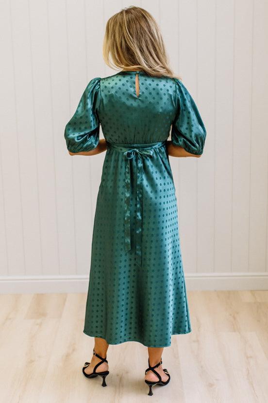 A woman viewed from behind wearing a teal green midi dress with a small polka dot pattern, puffed short sleeves, and a tie waist, paired with black strappy heels against a white paneled wall and light wood floor.