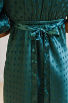 A woman modeling a teal green midi-length slip dress with a black polka dot pattern, black lace trim at the V-neck, and puff sleeves, paired with black heels, standing in a room with white paneled walls and light wood floors.