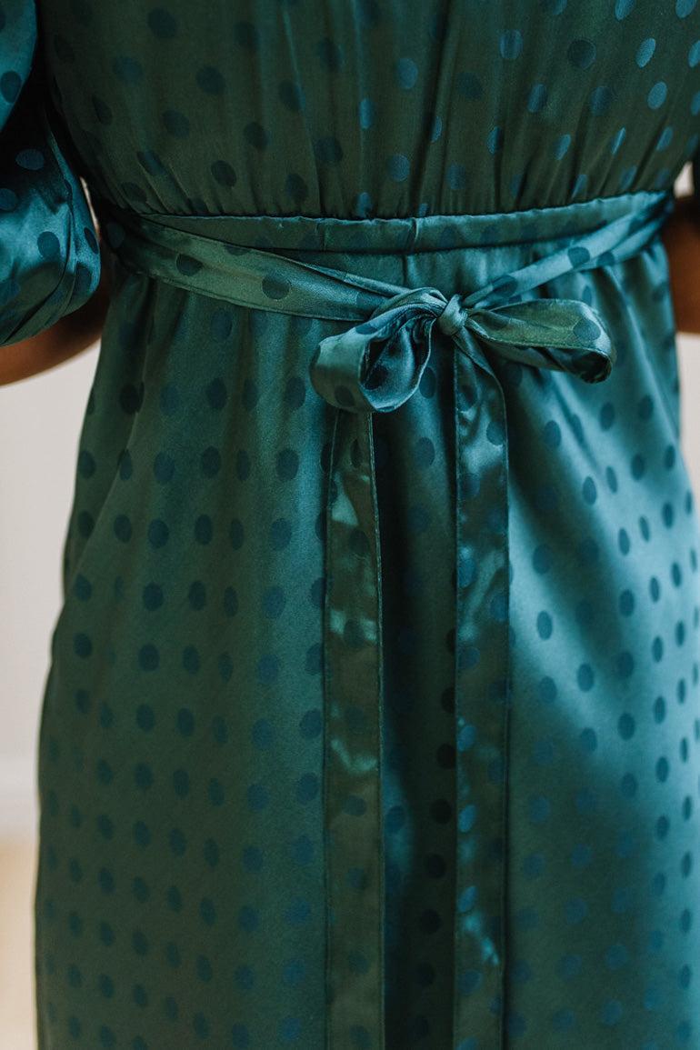 A woman modeling a teal green midi-length slip dress with a black polka dot pattern, black lace trim at the V-neck, and puff sleeves, paired with black heels, standing in a room with white paneled walls and light wood floors.