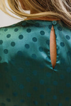 A woman modeling a teal green midi-length slip dress with a black polka dot pattern, black lace trim at the V-neck, and puff sleeves, paired with black heels, standing in a room with white paneled walls and light wood floors.