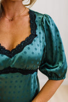 A woman modeling a teal green midi-length slip dress with a black polka dot pattern, black lace trim at the V-neck, and puff sleeves, paired with black heels, standing in a room with white paneled walls and light wood floors.