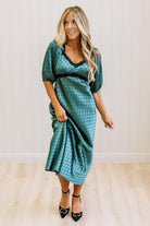A woman modeling a teal green midi-length slip dress with a black polka dot pattern, black lace trim at the V-neck, and puff sleeves, paired with black heels, standing in a room with white paneled walls and light wood floors.