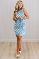 Woman wearing a blue dress with white polka dots against a light wooden floor and white wall background.