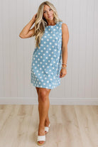 Woman wearing a blue dress with white polka dots against a light wooden floor and white wall background.