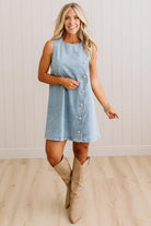A woman modeling a light blue sleeveless mini dress with a button-down front, paired with beige knee-high boots, standing in front of a white paneled wall and light wood floor.