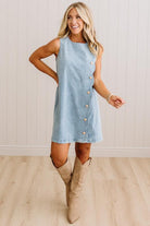 A woman modeling a light blue sleeveless denim dress with a row of gold buttons down the front, paired with beige knee-high boots, standing in front of a white paneled wall and light wood floor.