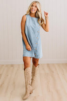 A woman modeling a sleeveless light blue denim dress with a row of decorative buttons down the front, paired with beige cowboy boots, standing in front of a white paneled wall and light wood floor.