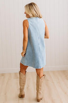 A woman shown from behind wearing a sleeveless light blue denim dress and beige cowboy boots, standing in a room with light wood floors and white paneled walls.