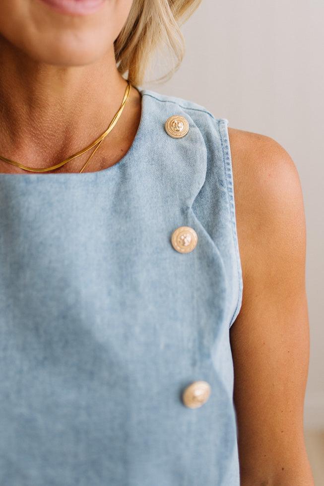 Close-up of a woman wearing a light blue sleeveless top with three decorative gold buttons on the front, paired with a thin gold necklace.