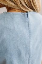 Close-up back view of a person wearing a light blue denim or chambray top, showing the neckline, a thin gold chain necklace, and blonde hair.