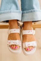 White sandals with floral straps worn with light blue jeans on a neutral background