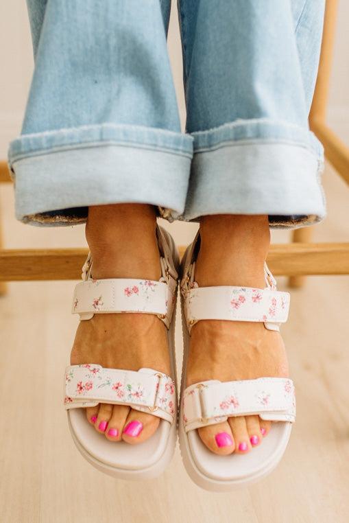 White sandals with floral straps worn with rolled-up denim jeans on a neutral background