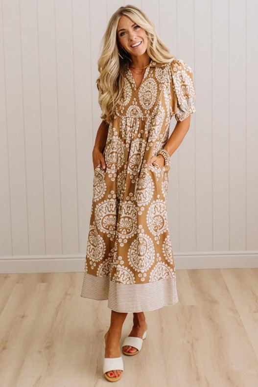 A woman modeling a tan and white paisley midi dress with puff sleeves and a striped hem, standing in a room with light wood floors and white paneled walls.