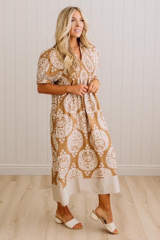 A woman modeling a tan and white patterned midi dress with short sleeves and a textured hem, standing in front of a white paneled wall and light wood floor.