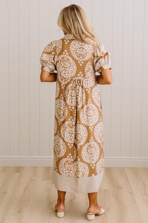 A woman viewed from behind wearing a tan and white patterned midi dress with puff sleeves and a tie waist, standing in front of a white paneled wall and light wood floor.
