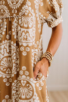Close-up of a person wearing a brown and white paisley patterned dress, with a hand in a pocket and stacked gold-toned bracelets visible against a soft neutral background.
