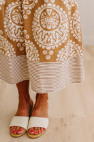 A close-up of a person wearing a brown and cream patterned maxi skirt with a striped hem, paired with white slide sandals on a light wood floor.