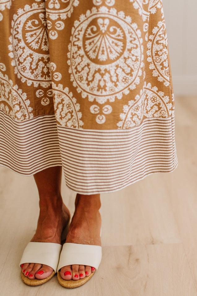 A close-up of a person wearing a brown and cream patterned maxi skirt with a striped hem, paired with white slide sandals on a light wood floor.