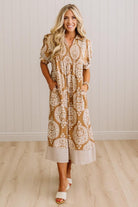 A woman modeling a tan and white patterned midi dress with puff sleeves and side pockets, standing in a room with light wood floors and white paneled walls.