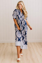 A woman modeling a navy blue and white paisley midi dress with a striped hem, standing in a room with light wood floors and white paneled walls.